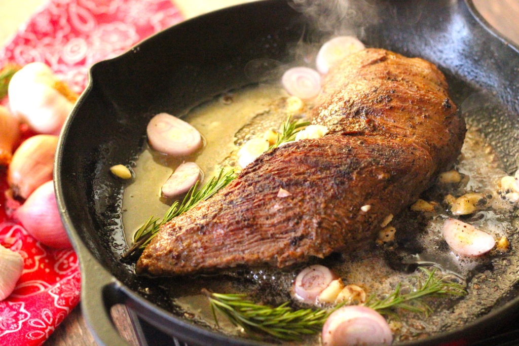Tri tip roast cooking in a cast iron skillet with shallots, garlic, butter and rosemary.