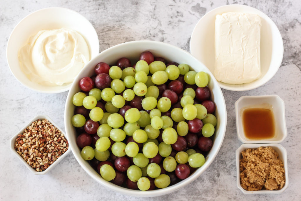 ingredients needed to make grape salad. Sour cream, cream cheese, pecans, red and green grapes, vanilla and brown sugar in white bowls.