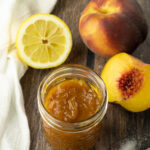 peach jam in a glass jar surrounded by peaches and a sliced lemon