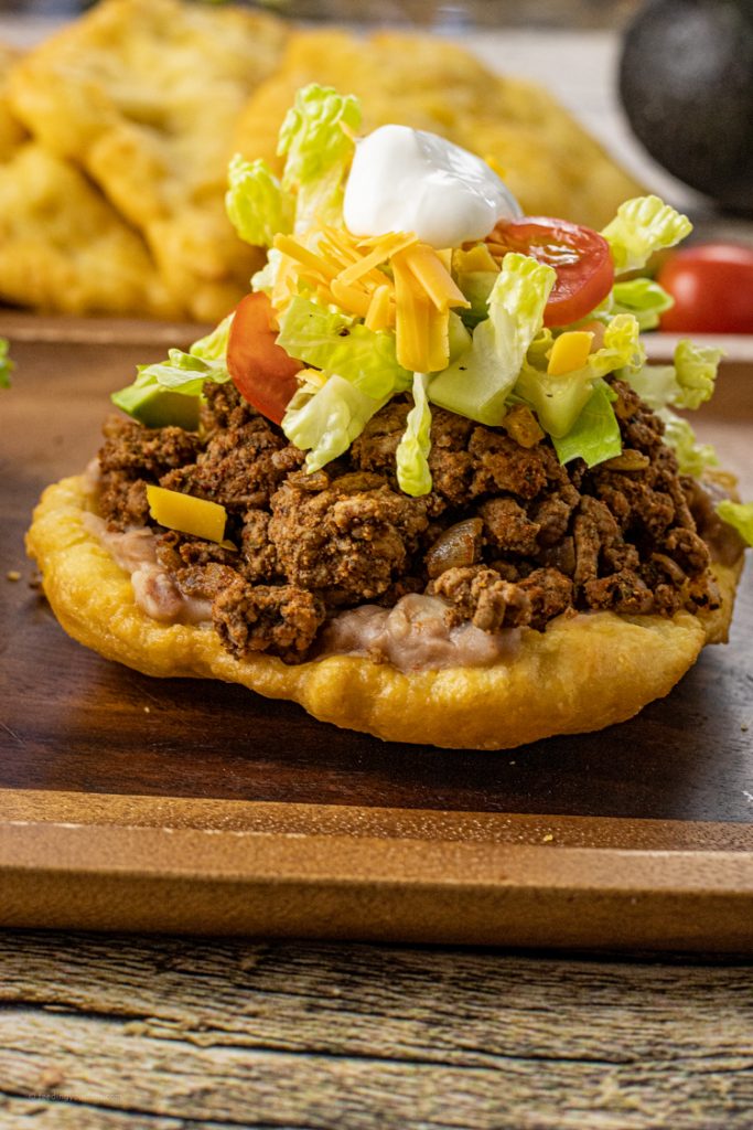 fry bread taco recipe topped with beans, meat, lettuce, cheese and sour cream