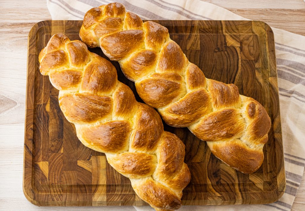two loaves of baked, braided bread on a wooden cutting board