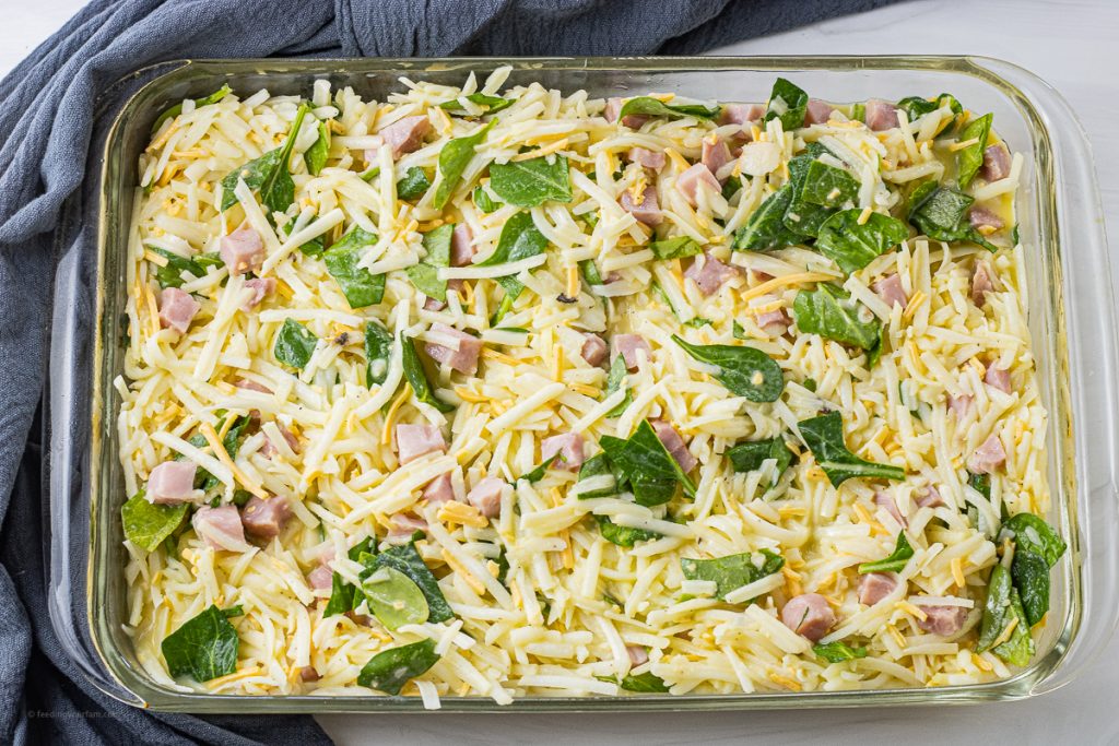 uncooked egg casserole with ham, hash browns, cheese and spinach in a glass baking dish