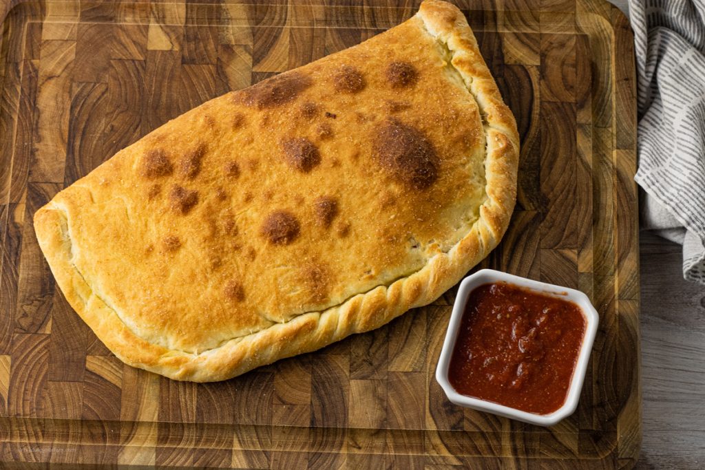 browned calzone on a wooden cutting board with a small bowl of pizza sauce