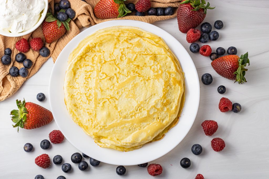 pile of crepes on a white plate surrounded by berries