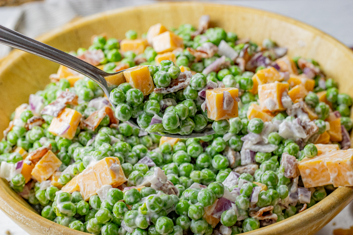 spoon scooping out some green pea salad with cheese and bacon in a creamy dressing from a wooden bowl