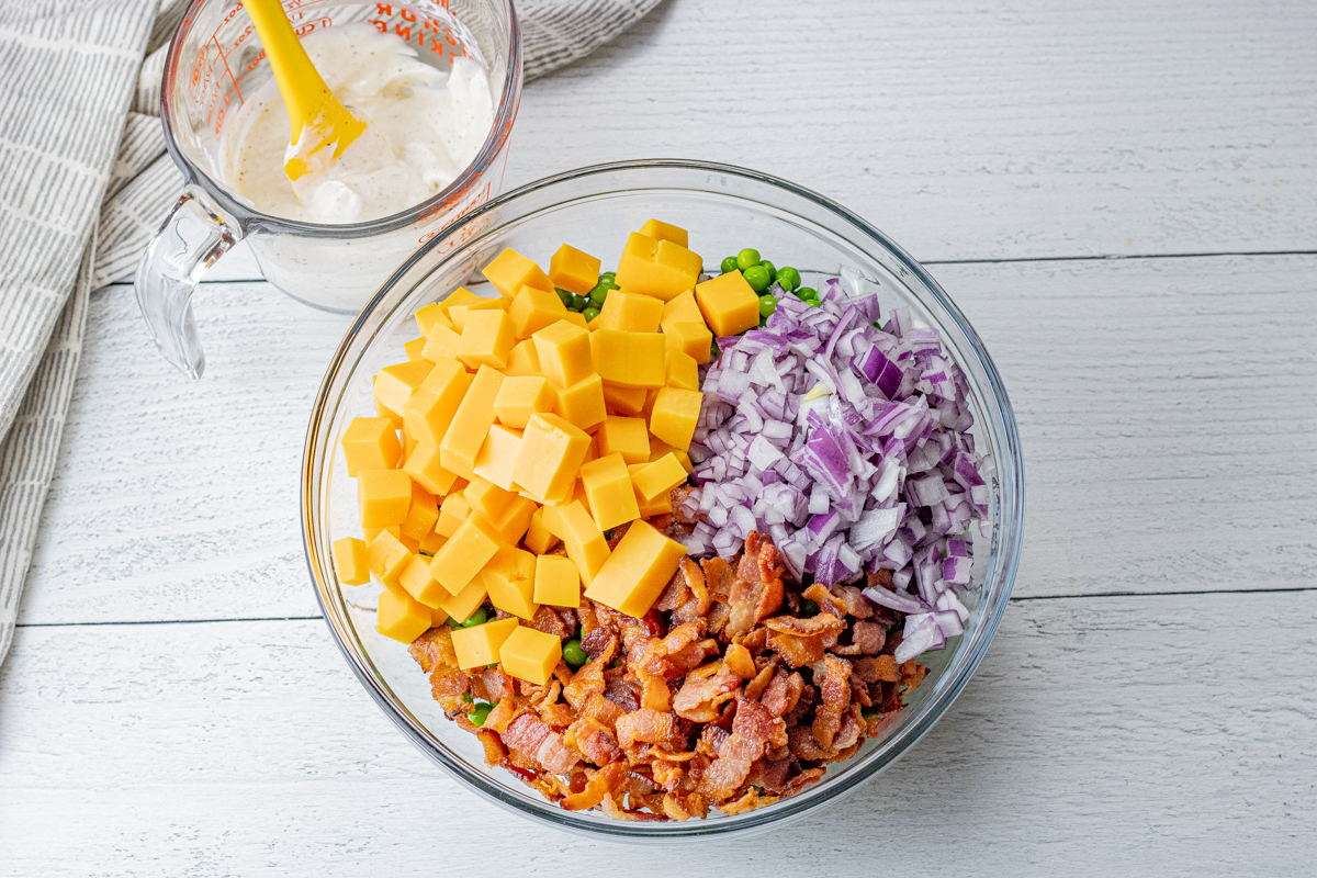 cheese cubes, red onion, crisp bacon and green peas in a glass bowl with a cup of creamy dressing in a measuring cup next to it