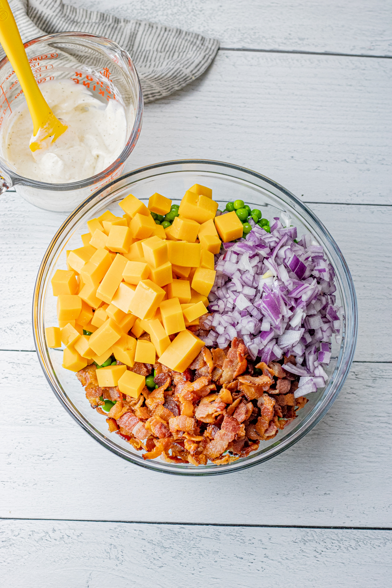 cheese cubes, red onion, crisp bacon and green peas in a glass bowl with a cup of creamy dressing in a measuring cup next to it