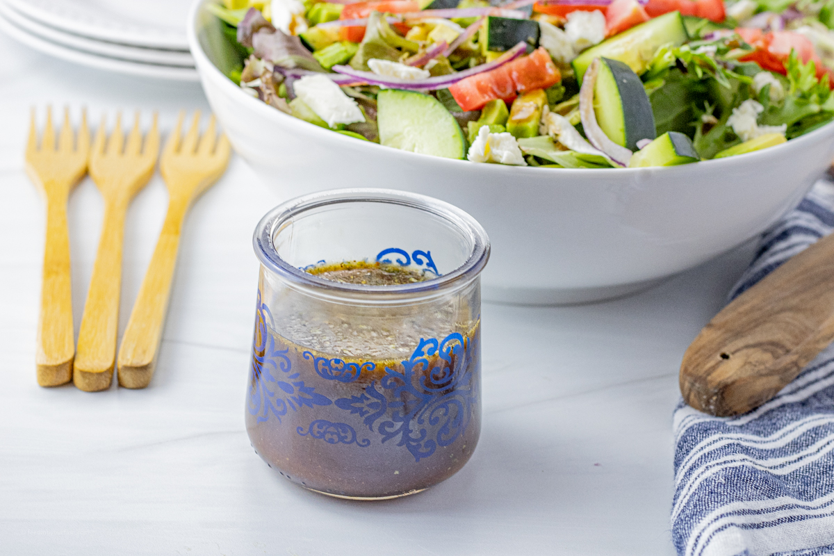 small glass jar with balsamic vinegar dressing in it in front of a large white bowl filled with green salad