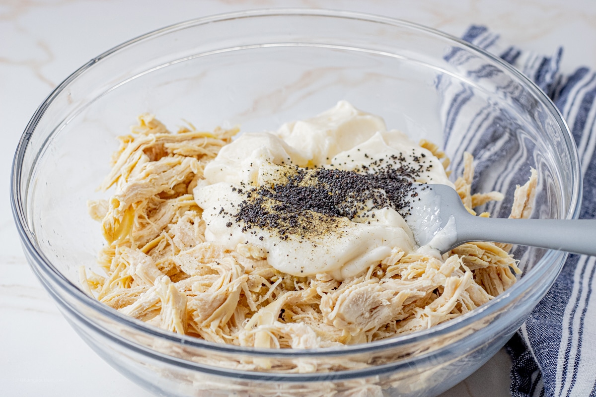 shredded chicken topped with mayonnaise, salt, pepper and poppyseeds all in a glass mixing bowl