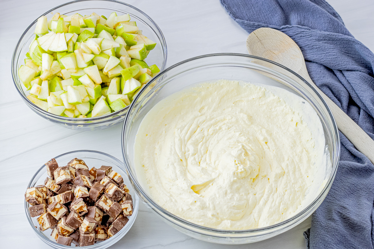 Three mixing bowls, one filled with vanilla pudding mixture, chopped green apples and chopped snickers candy bars