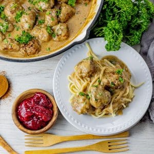 meatballs in a brown gravy on a white plate with a small wooden bowl of raspberry jam next to it