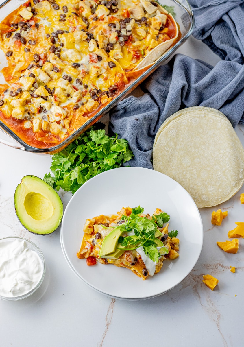 a pan filled with a layered tortilla casserole alongside a white plate with a slice of the casserole on it