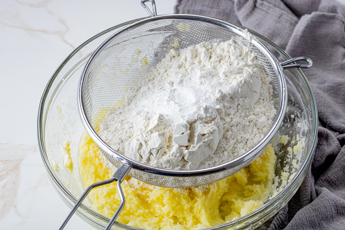 flour in a mesh strainer over creamed butter and sugar in a glass mixing bowl
