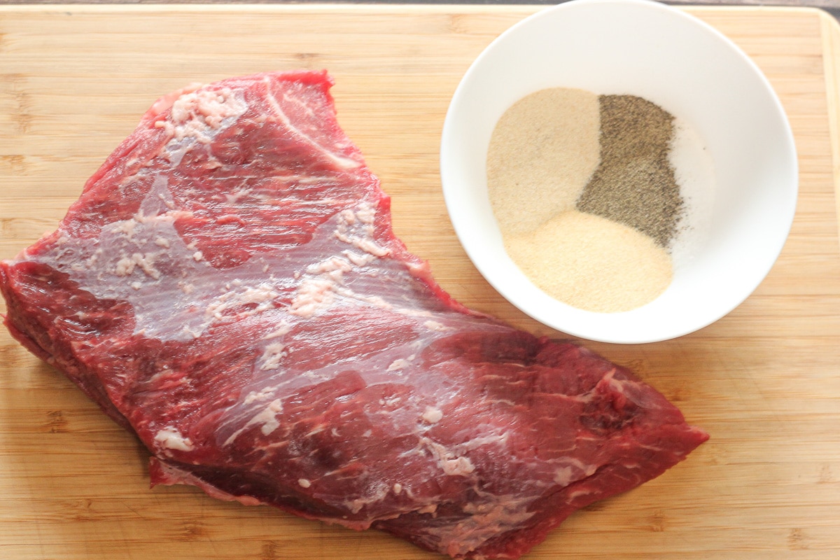uncooked tri tip beef roast next to a small bowl of spices on a wooden cutting board