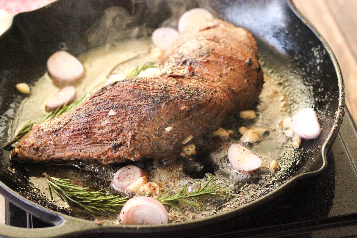 beef tri tip roast cooking in a cast iron pan with shallots, rosemary and garlic