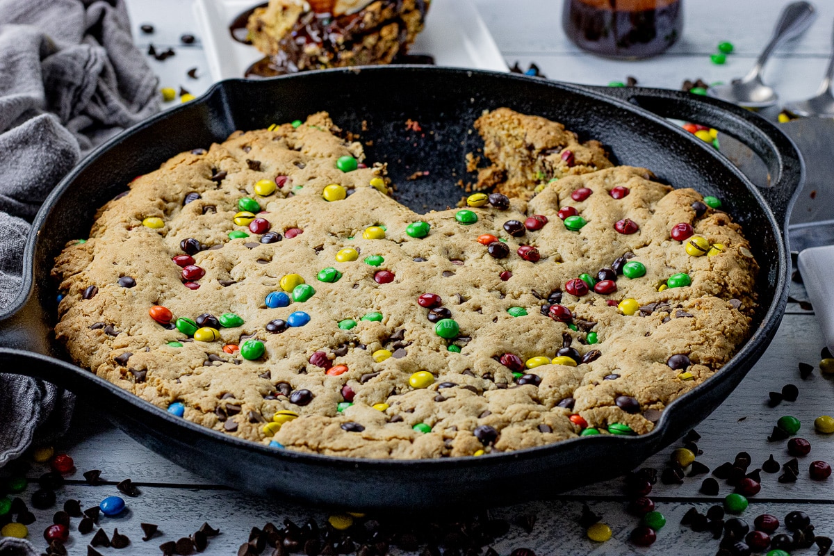 cast iron pan filled with a cookie with chocolate chips and m and ms with a piece cut out