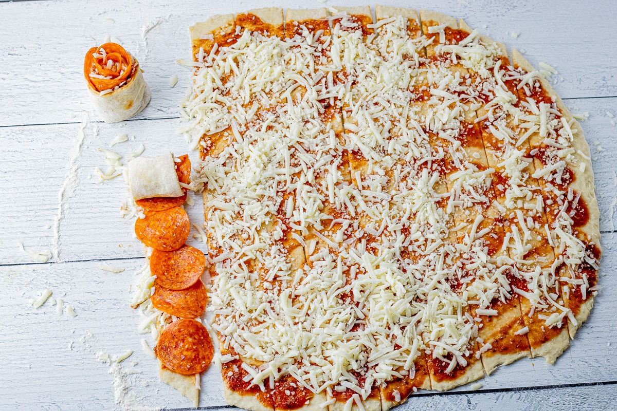 pizza dough topped with shredded cheese, and pepperonis being rolled up into a roll