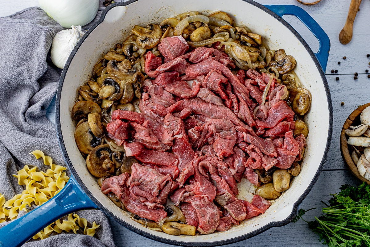 uncooked strips of steak in a cast iron pan with cooked mushrooms and onions