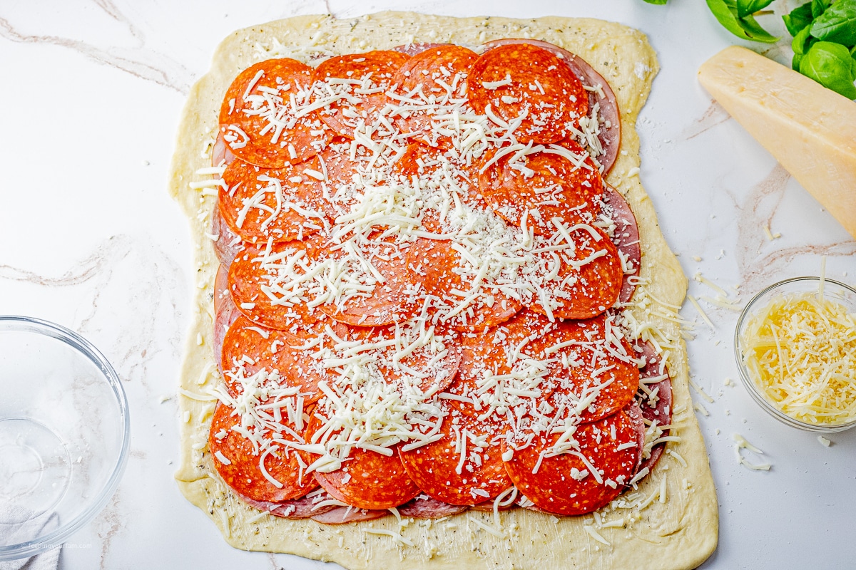 layers of ham, salami, pepperoni and cheese on a rectangle of pizza dough