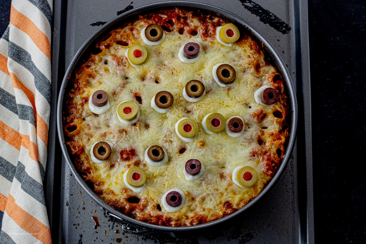 baked rigatoni in a round springform pan, topped with melted cheese and small mozzarella pearls, then topped with olives to look like eyeballs