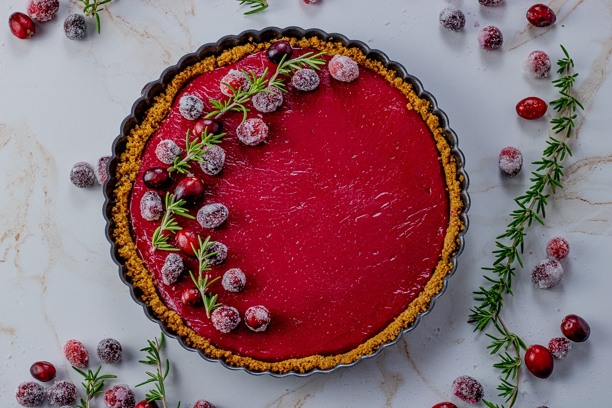 red, cranberry tart with a graham cracker crust in a tart pan. The tart is topped with sugared cranberries and rosemary sprigs