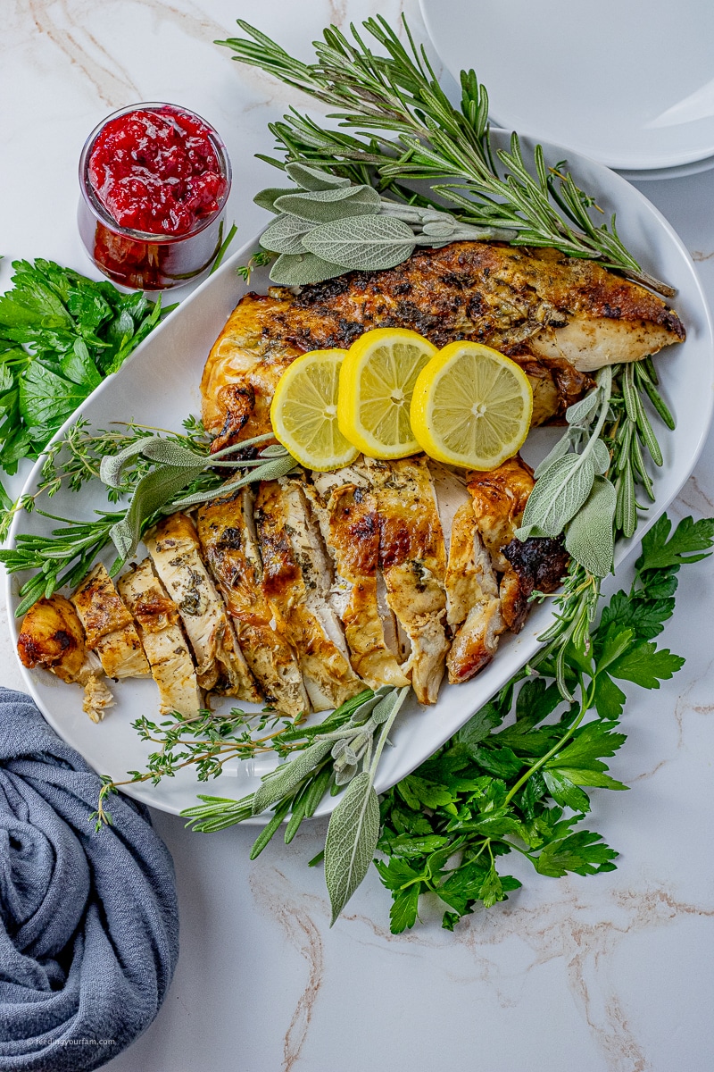 two turkey breasts on a white serving platter with fresh sprigs of rosemary, time and sage