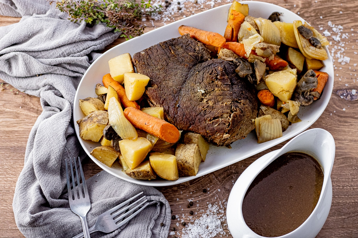 cooked pot roast on a white platter surrounded by carrots, potatoes and onions