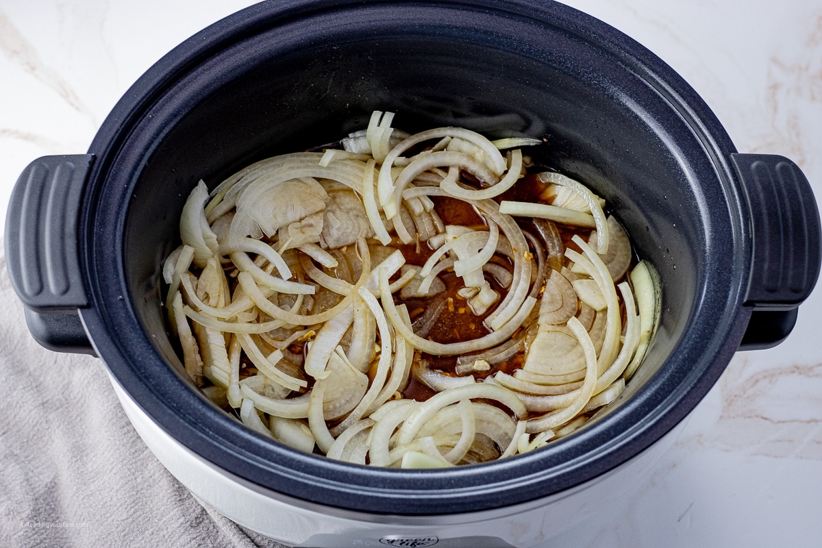 sliced onions on top of a chuck roast in the pot of a slow cooker