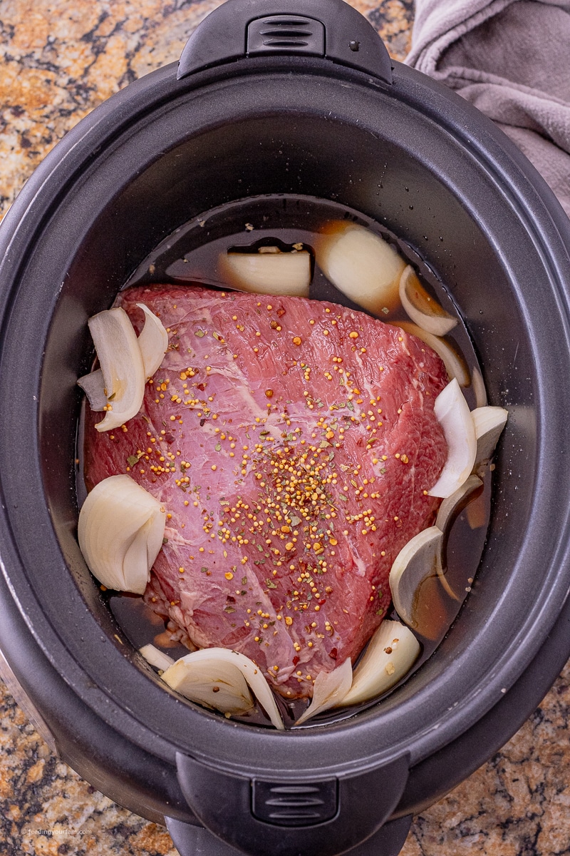 uncooked corned beef in a slow cooker with sliced onions and beef broth around it