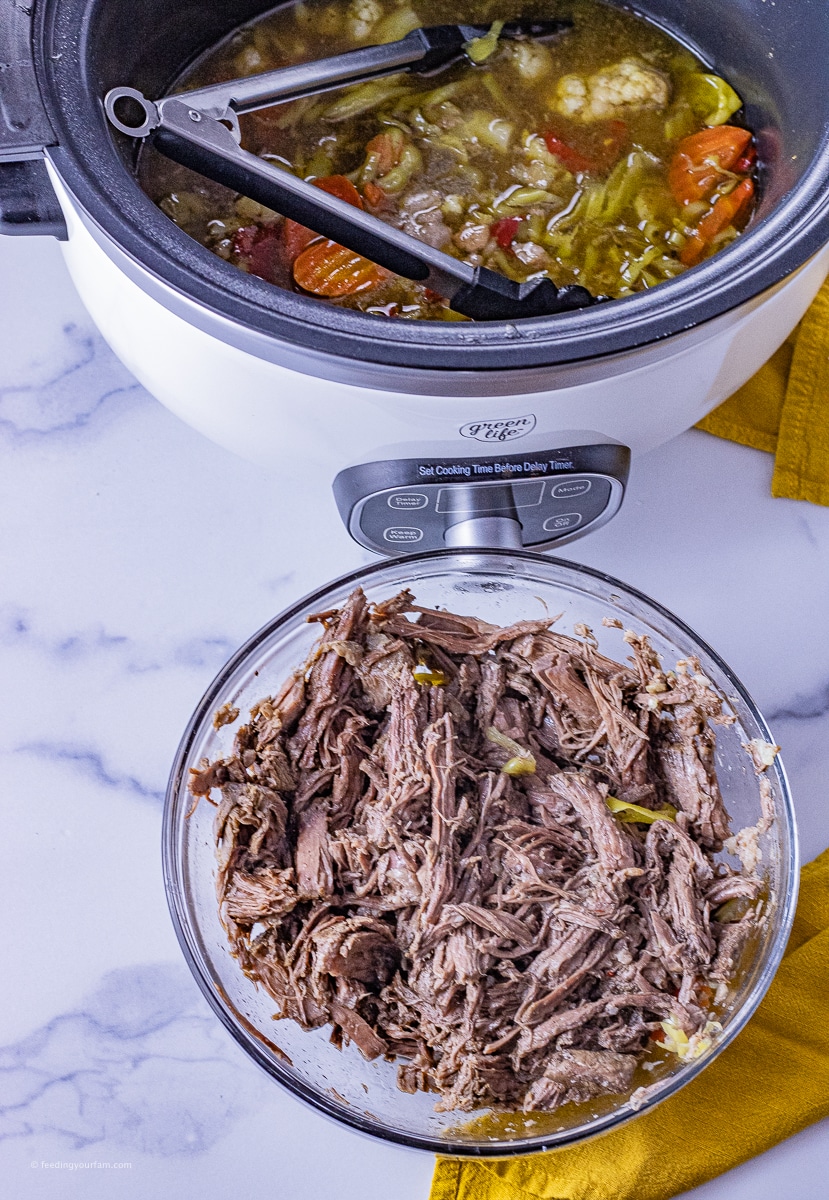 shredded beef in a bowl with a slow cooker filled with vegetables behind it