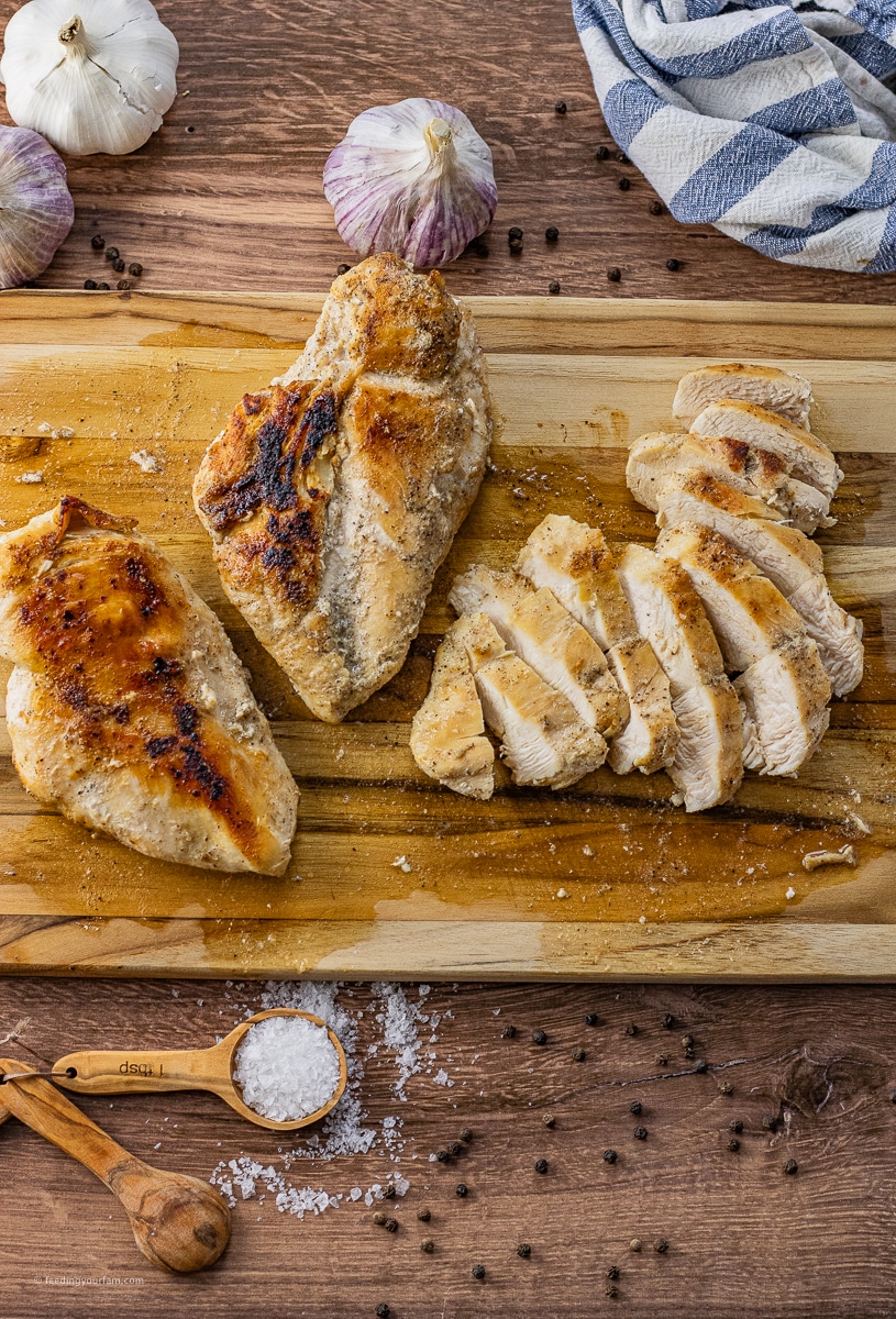 three chicken breasts on a wooden cutting board, one is sliced. They are cooked chicken