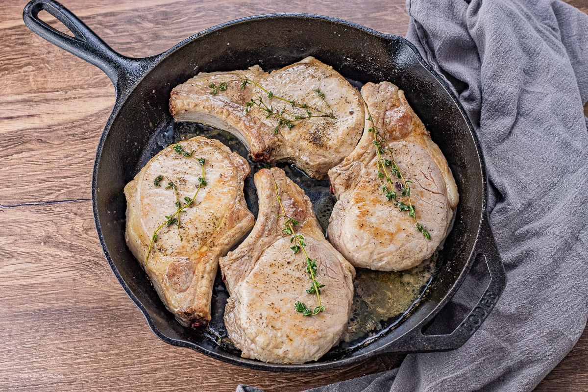 four bone in pork chops cooking in a cast iron pan with sprigs of thyme