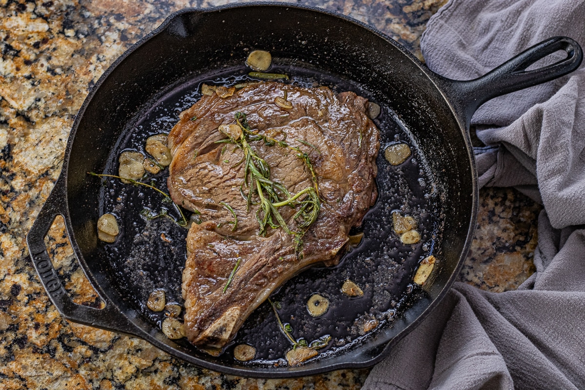 ribeye steak cooking in a cast iron pan with rosemary, thyme and sliced garlic