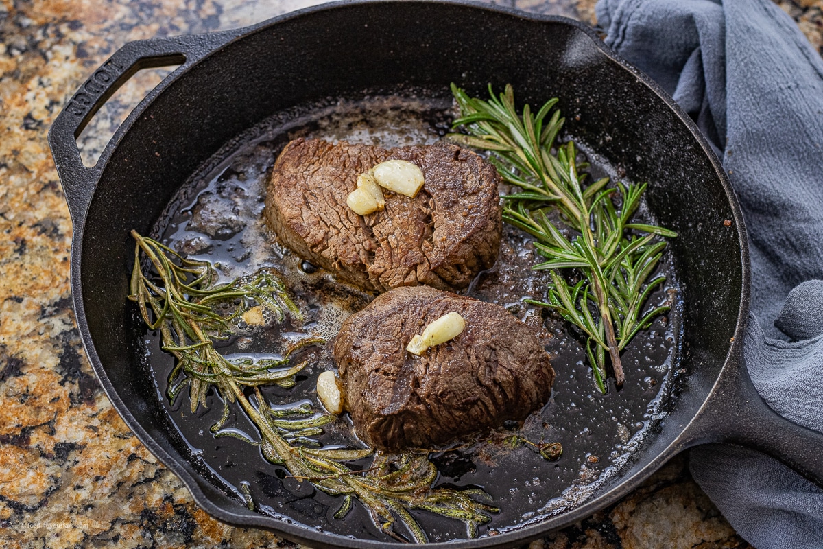 two filet mignons topped with garlic cloves in a cast iron pan with rosemary sprigs