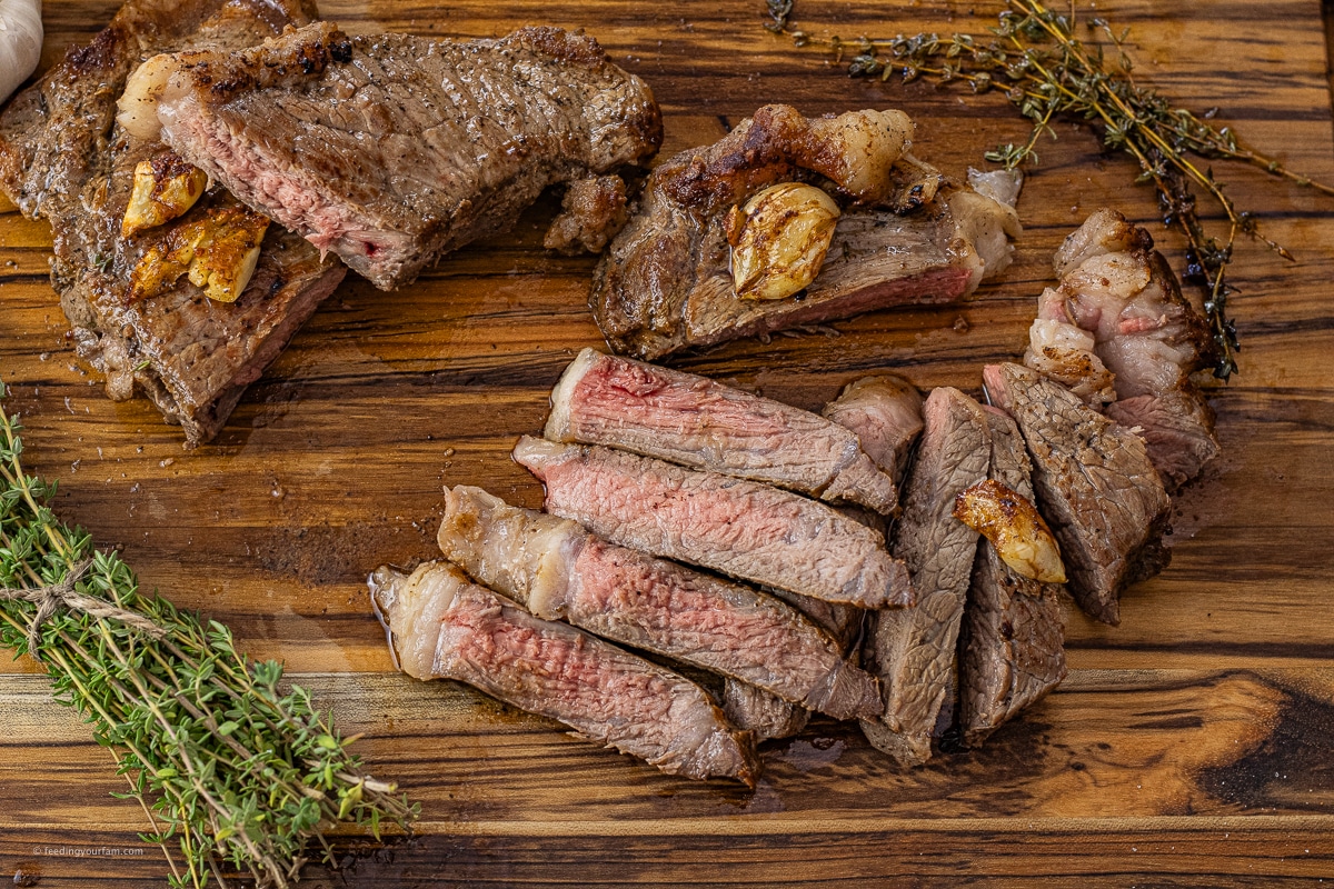 cooked and sliced new york steak on a wooden cutting board with thyme sprigs and cooked garlic