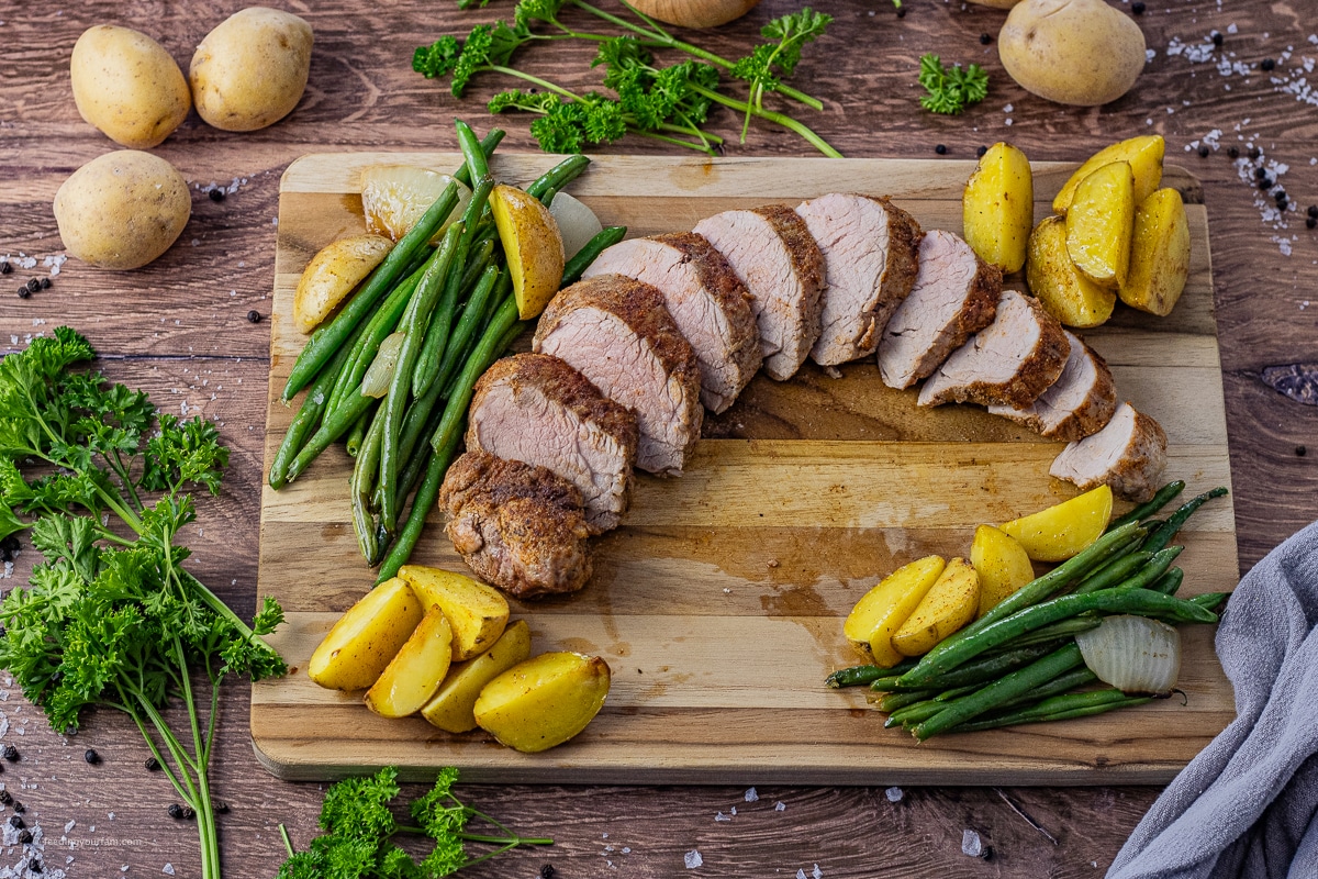 cooked pork tenderloin, sliced and on a wooden cutting board with green beans and potatoes