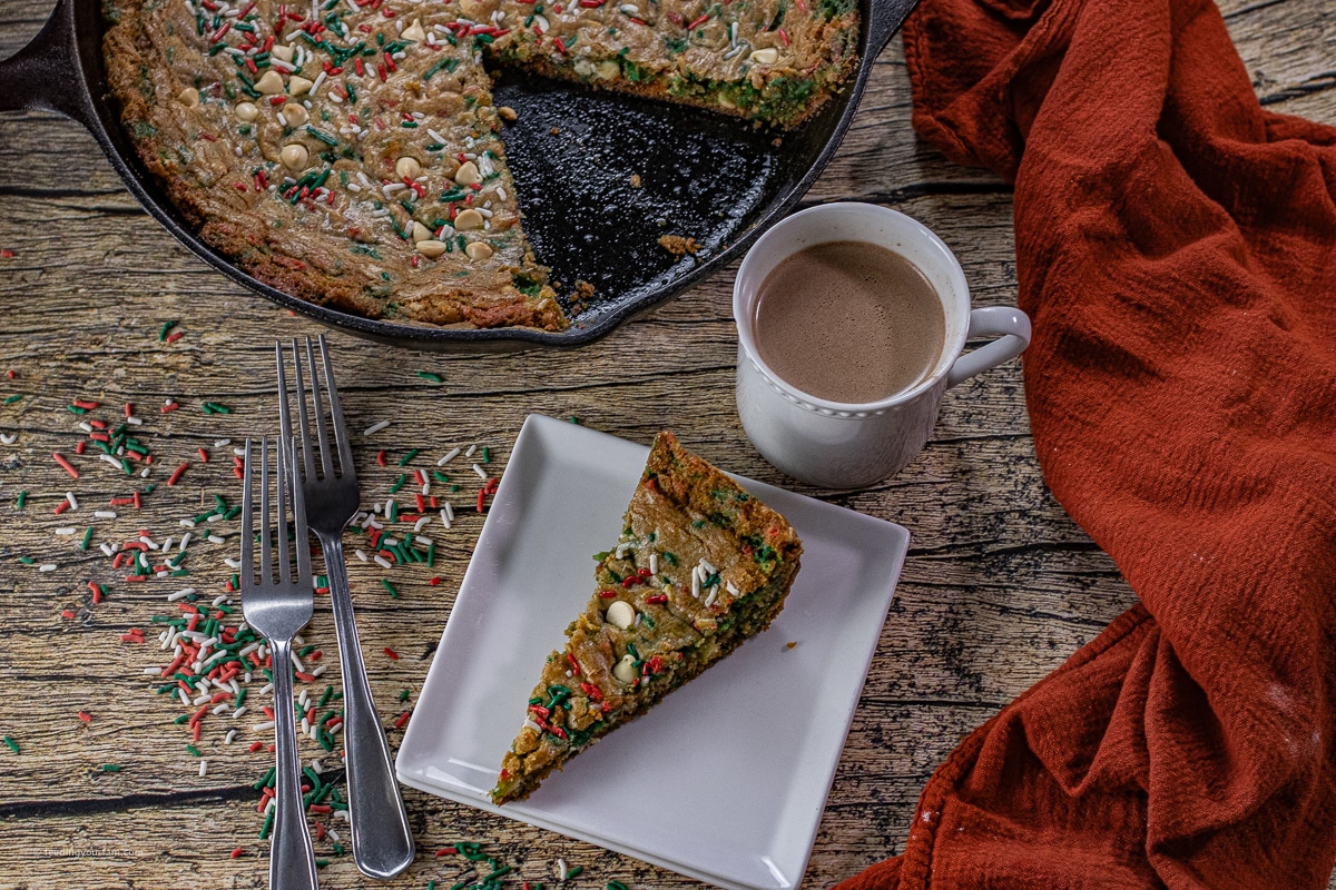 slice of Christmas sprinkle skillet cookie on a white plate next to a mug of hot chocolate