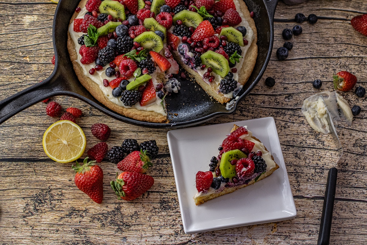 slice of fruit pizza on a white, square plate next to a cast iron skillet with the remaining fruit pizza inside