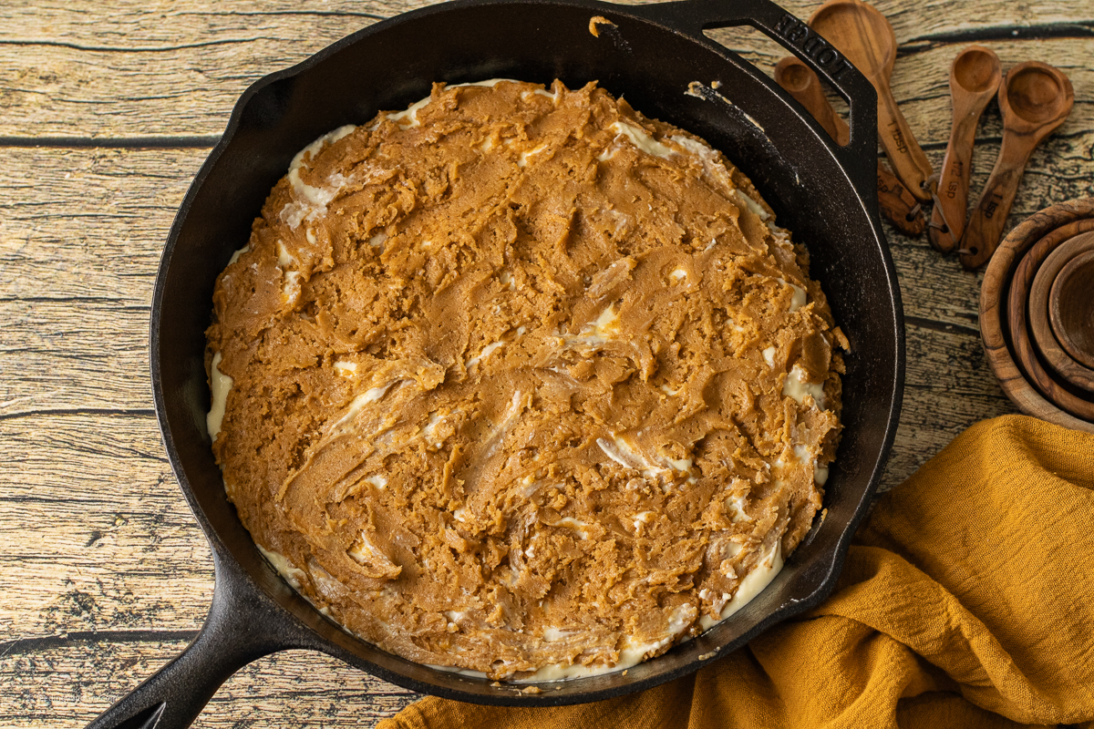 A cast iron skillet filled with a golden pumpkin cookie dough swirled with a creamy cheesecake layer uncooked